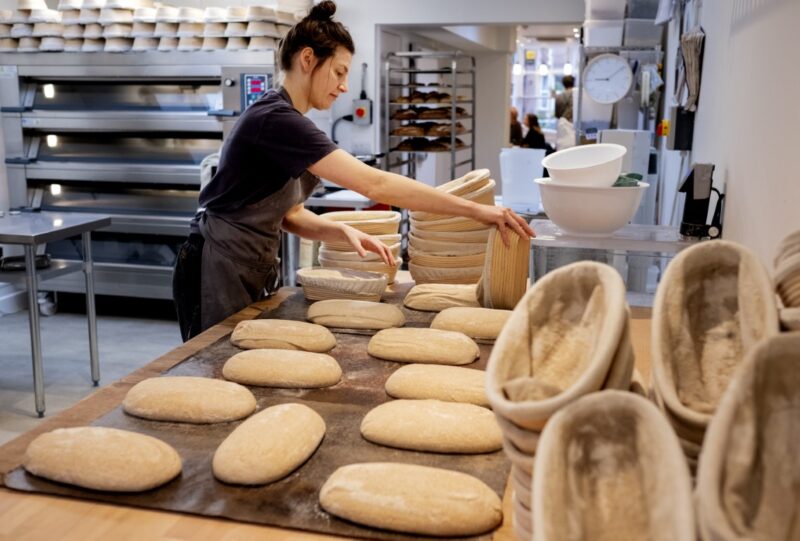 Woman wearing apron standing in an artisan bakery, shaping sourdough loaves for baking.