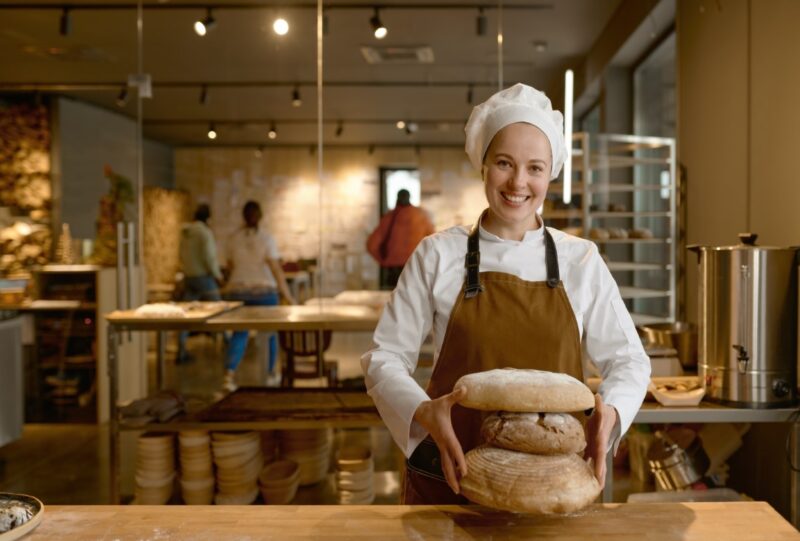 Portrait of happy baker holding stack of freshly baked bread. Young female chef proud of making natural organic pastry standing at table and looking at camera